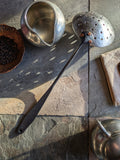 (Hand-forged stainless steel slotted spoon with a black-forged handle and polished perforated bowl showing back side.)