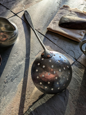 (Hand-forged stainless steel slotted spoon with a black-forged handle and polished perforated bowl.)