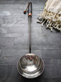 Full view of a hand-forged sauna water ladle with polished hammered stainless steel bowl and black ash handle, handmade in Quebec.