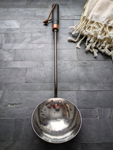Full view of a hand-forged sauna water ladle with polished hammered stainless steel bowl and black ash handle, handmade in Quebec.