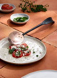 full view of hand forged stainless steel and copper serving spoon resting on a plate of tomatoes
